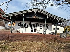 zu sehen ist die Hütte in der Vorderansicht, mit zwei Fenstern und einer Türe. Witterungsbedingte Abnutzung am Holz.