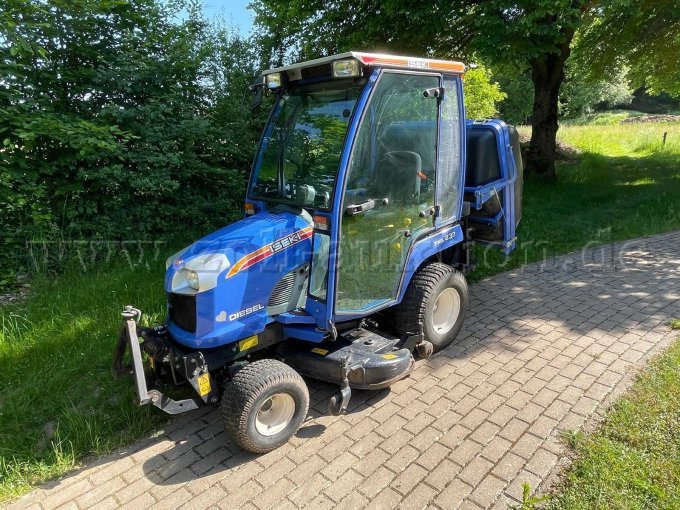 Zu sehen ist ein blauer ISEKI TXG 237A Kompakttraktor von vorne rechts. Hinten am Fahrzeug erkennt man eine Grasaufnahme und unter dem Fahrzeug das Mähwerk.