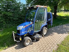 Zu sehen ist ein blauer ISEKI TXG 237A Kompakttraktor von vorne rechts. Hinten am Fahrzeug erkennt man eine Grasaufnahme und unter dem Fahrzeug das Mähwerk.