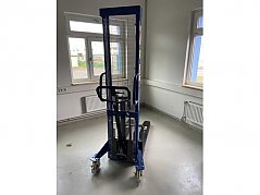 Frontalansicht des Hochhubwagens im Profil von vorne. Gut zu erkennen sind das Hubgerüst mit Schutzgitter, die Deichsel, die blauen Ausleger mit den Lenkrollen und der Fußhebel.