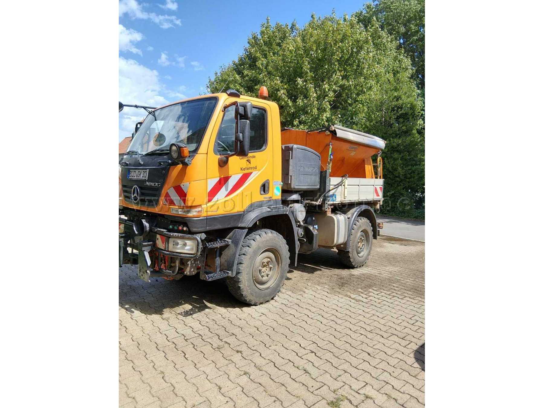 Außenansicht des Fahrzeugs von vorne links. Zu sehen sind Fahrerkabine, Bereifung, Pritschenbereich und der aufgebaute Winterdienst-Streuer.