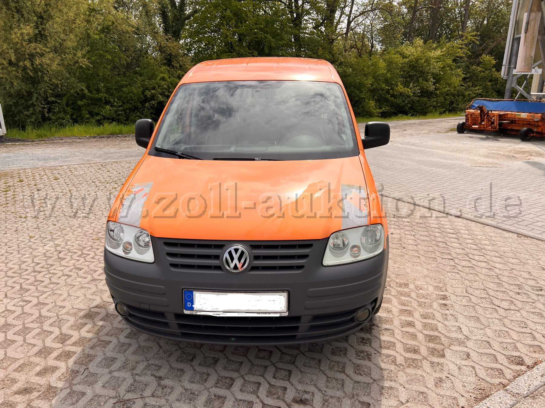 Auf dem Bild erkennt man die Front eines orangefarbenen Kommunalfahrzeugs. Der Caddy hat auf der Motorhaube links und rechts zwei bereits verblasste und beschädigte Warnmarkierungen.